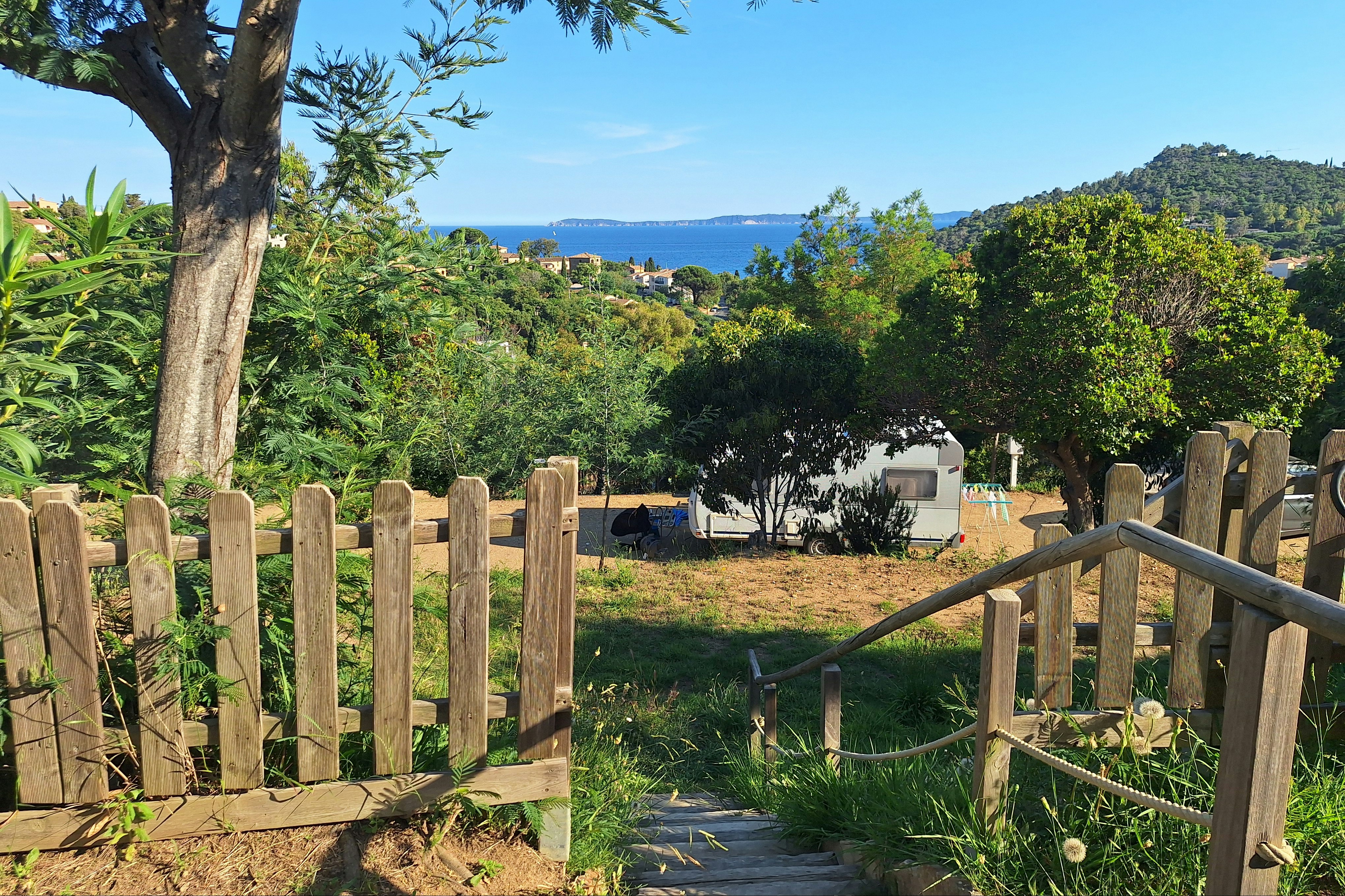 Wohnwagen auf einem Campingplatz, umgeben von Bäumen und einem Holzlattenzaun, mit Blick auf das Meer in der Ferne.