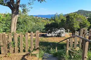 Wohnwagen auf einem Campingplatz, umgeben von Bäumen und einem Holzlattenzaun, mit Blick auf das Meer in der Ferne.