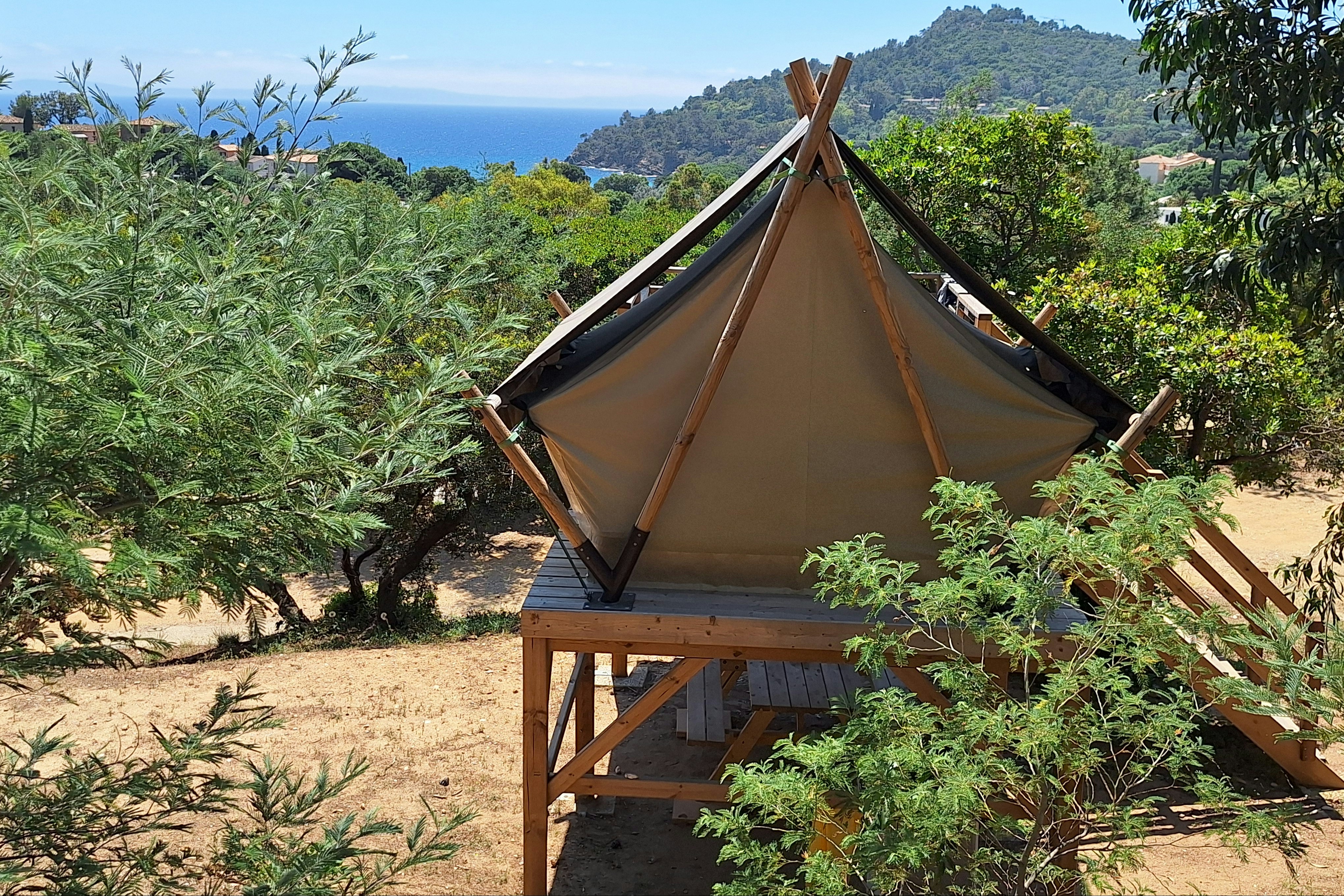 Zelt auf einer Holzplattform im Campingbereich, umgeben von Büschen und Bäumen, mit Blick auf das Meer und einen bewaldeten Hügel.