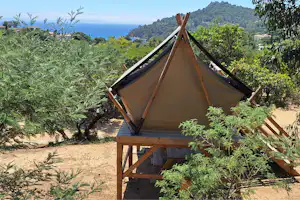 Zelt auf einer Holzplattform im Campingbereich, umgeben von Büschen und Bäumen, mit Blick auf das Meer und einen bewaldeten Hügel.