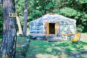 Yurt-Zelt mit geöffnetem Eingang auf einem Stellplatz, Holzpodest und Liegestuhl, umgeben von Bäumen im Campinggrün.