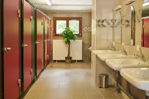 Badezimmer im Campingbereich mit mehreren Waschbecken, Spiegeln, Fliesenboden und roten Türen im Flur.
