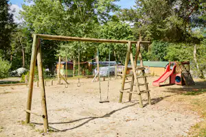 Spielplatz mit Holzschaukel und roter Rutsche auf sandigem Boden, im Hintergrund Bäume und Campinggebäude mit grünem Dach.