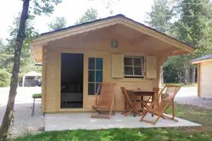 Holzbungalow mit Terrasse, zwei Liegestühlen und Tisch vor großen Fenstern, umgeben von Bäumen und Gartenweg.