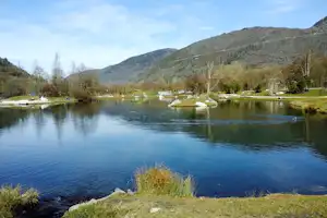Ruhiger Campingsee mit Steinen am Ufer, Wasserpflanzen und Booten, im Hintergrund bewaldete Berge und Bäume.