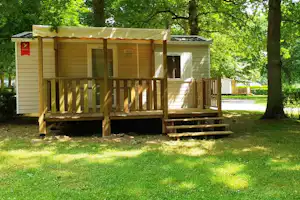Mobilheim mit Holzveranda und Stufen, umgeben von Rasen und Bäumen im Campingbereich.