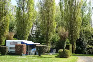 Wohnwagen mit Vorzelt und Sitzgruppe auf einem Campingplatz, umgeben von Hecken, Rasen und hohen Bäumen.