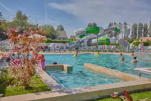 Schwimmbecken auf dem Campingplatz mit grünen und weißen Rutschen, Liegen und Blumenbeeten am Beckenrand.