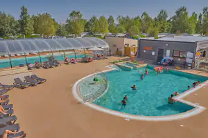 Großes Camping-Schwimmbecken mit mehreren Liegestühlen, Sonnenschirmen und Menschen im Wasser, daneben Gebäude und Bäume.