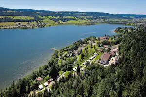 Luftaufnahme eines Campingplatzes am See mit Wohnwagen, Wegen und Häusern am Waldrand, umgeben von grünen Hügeln.