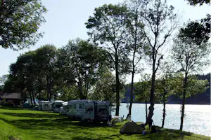 Reihe von Wohnmobilen auf einem Campingplatz am See, mit Zelt, Rasen und Bäumen am Ufer.