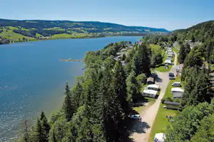 Campingplatz am See mit Wohnwagen und Stellplätzen, schmalem Weg, viel Grün und Wald, Blick von oben.