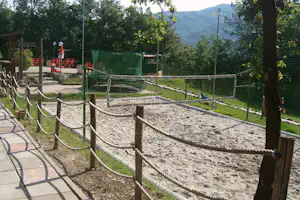Sandspielbereich mit Holzpfosten und Seilen, eingezäunt am Campingplatz, daneben Sitzreihen und Bäume mit Bergen im Hintergrund.