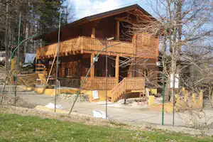 Holzhaus mit Veranda und Geländer, eingezäunt im Campinggelände, umgeben von Bäumen und Wegen.