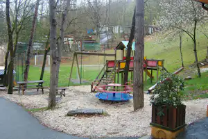 Spielplatz mit Schaukel und Rutsche auf Kiesfläche, Holzbänke und Tische zwischen Bäumen, im Hintergrund Zelte und Wege.