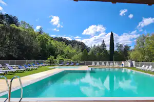 Camping-Pool mit grünem Rasen, blauen Liegen und weißen Sonnenliegen am Beckenrand, umgeben von Bäumen und Hügeln.