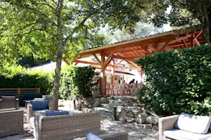 Holz-Terrasse am Campingplatz unter großen Bäumen, mit Stühlen, Steinumrandung und Sitzgelegenheiten im Garten.