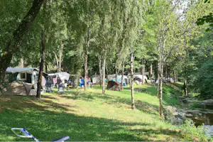 Campingplatz im grünen Wald mit mehreren Zelten, Grasfläche, Bäumen und einem kleinen Bachlauf am Rand.