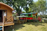 Camping Le Rouergue - Bauwagen- und Zirkuswagen-Mietunterkünfte mit überdachten Sitzmöglichkeiten im Halbschatten unter Bäumen