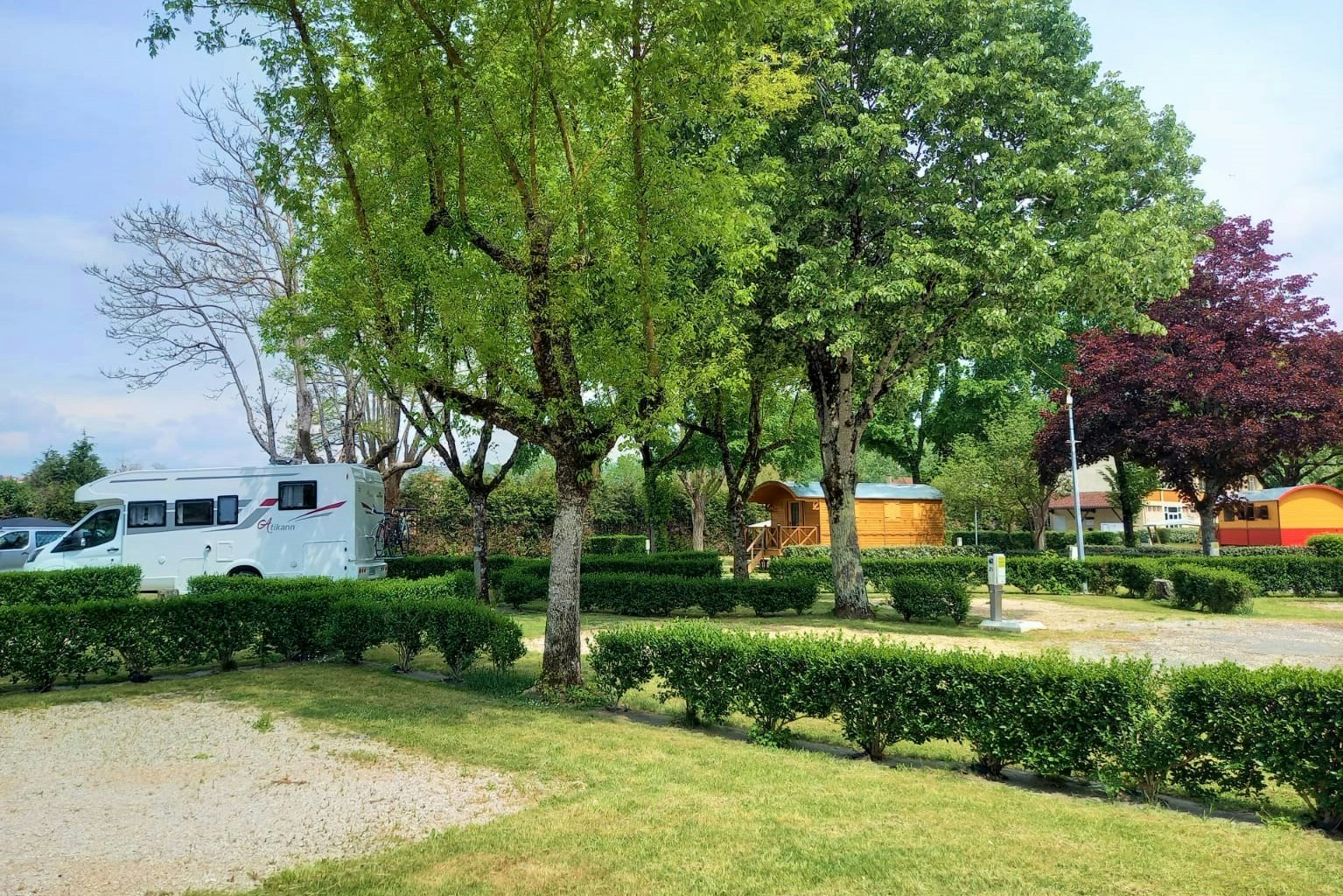 Camping Le Rouergue - Standplatz auf mit Gras durchwachsenem Sand umgeben von Wiese und durch Hecken abgetrennt mit Mietunterkünften im Hintergrund