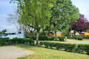 Camping Le Rouergue - Standplatz auf mit Gras durchwachsenem Sand umgeben von Wiese und durch Hecken abgetrennt mit Mietunterkünften im Hintergrund
