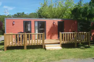 Bungalow mit Holzterrasse, roten Außenwänden und großen Fenstern, umgeben von Rasen und Bäumen.
