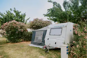 Wohnwagen mit Vorzelt auf einem Campingplatz, umgeben von Bäumen und blühenden Sträuchern im Garten.