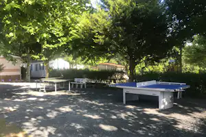 Campingplatz mit Bungalows, Tischtennisplatte und weißen Gartenmöbeln unter großen Bäumen im Schatten.
