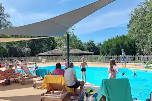 Camping-Pool mit Sonnensegel, Liegen und Tischen am Becken, im Hintergrund Bäume und ein Zaun.