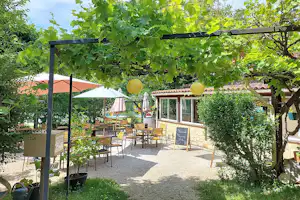 Terrasse mit Tischen und Stühlen unter Weinreben, daneben ein kleines Gebäude mit Fenstern und Steinmauer, im Grünen.