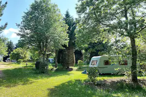 Wohnwagen auf einem Campingplatz im Grünen, umgeben von Bäumen und Hecken, mit Rasenfläche und kleinem Weg.