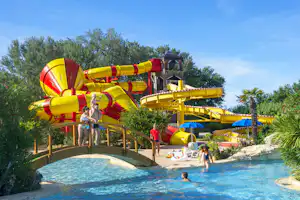 Großer Pool mit mehreren gelben Wasserrutschen und Sonnenschirmen, umgeben von Bäumen und Palmen auf dem Campingplatz.