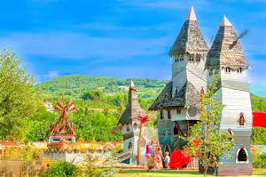 Märchenschloss mit Türmchen und bunten Fenstern vor grünen Hügeln, im Vordergrund Bäume und ein Spielbereich.