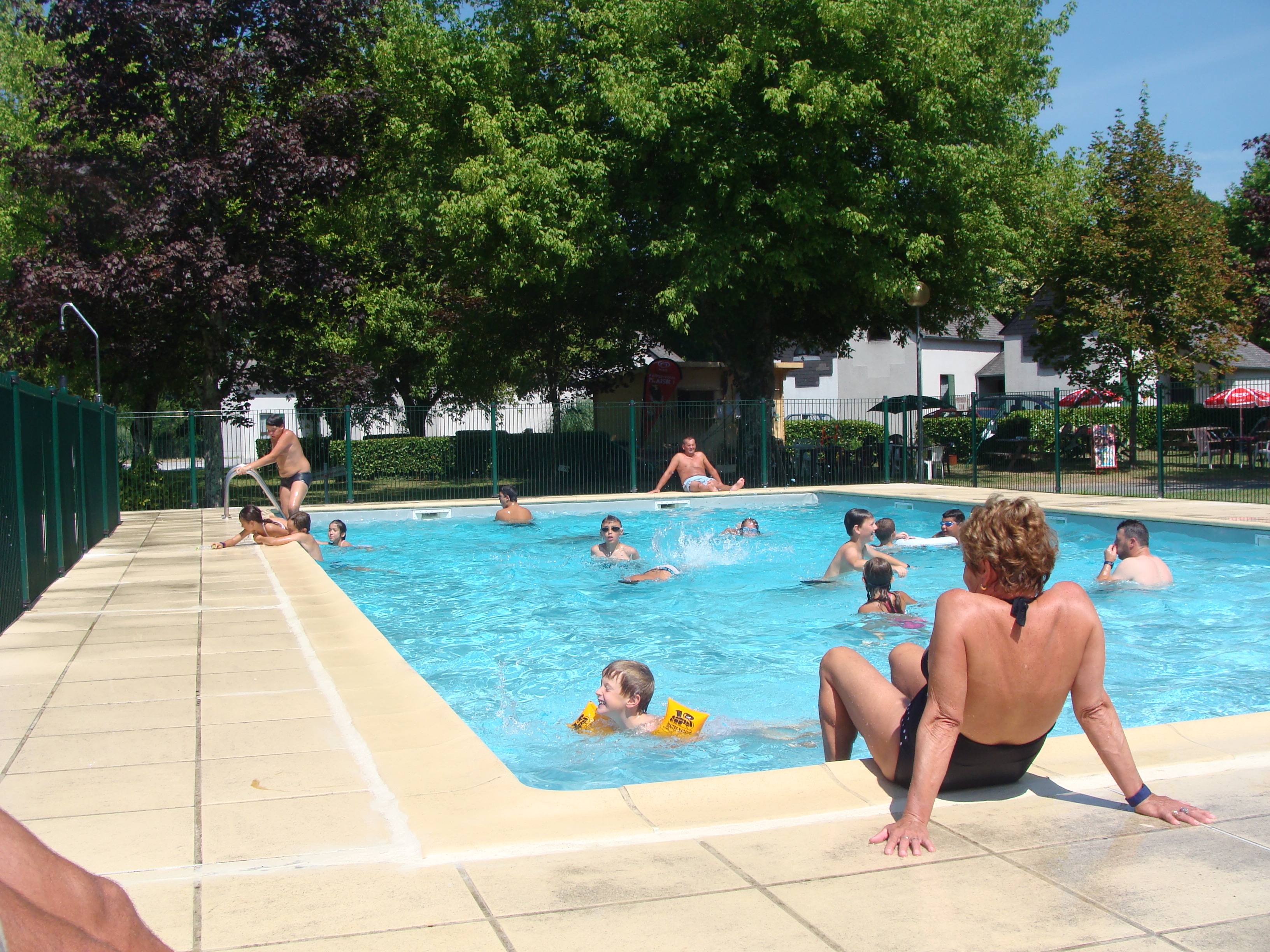 Schwimmbecken auf dem Campingplatz mit mehreren Gästen im Wasser, Sonnenschirme und Bäume im Hintergrund.