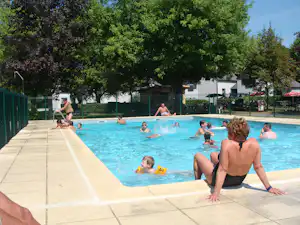 Schwimmbecken auf dem Campingplatz mit mehreren Gästen im Wasser, Sonnenschirme und Bäume im Hintergrund.