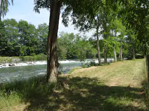 Fluss mit ruhigem Ufer und Grasfläche am Campingplatz, gesäumt von Bäumen und dichtem Grün.