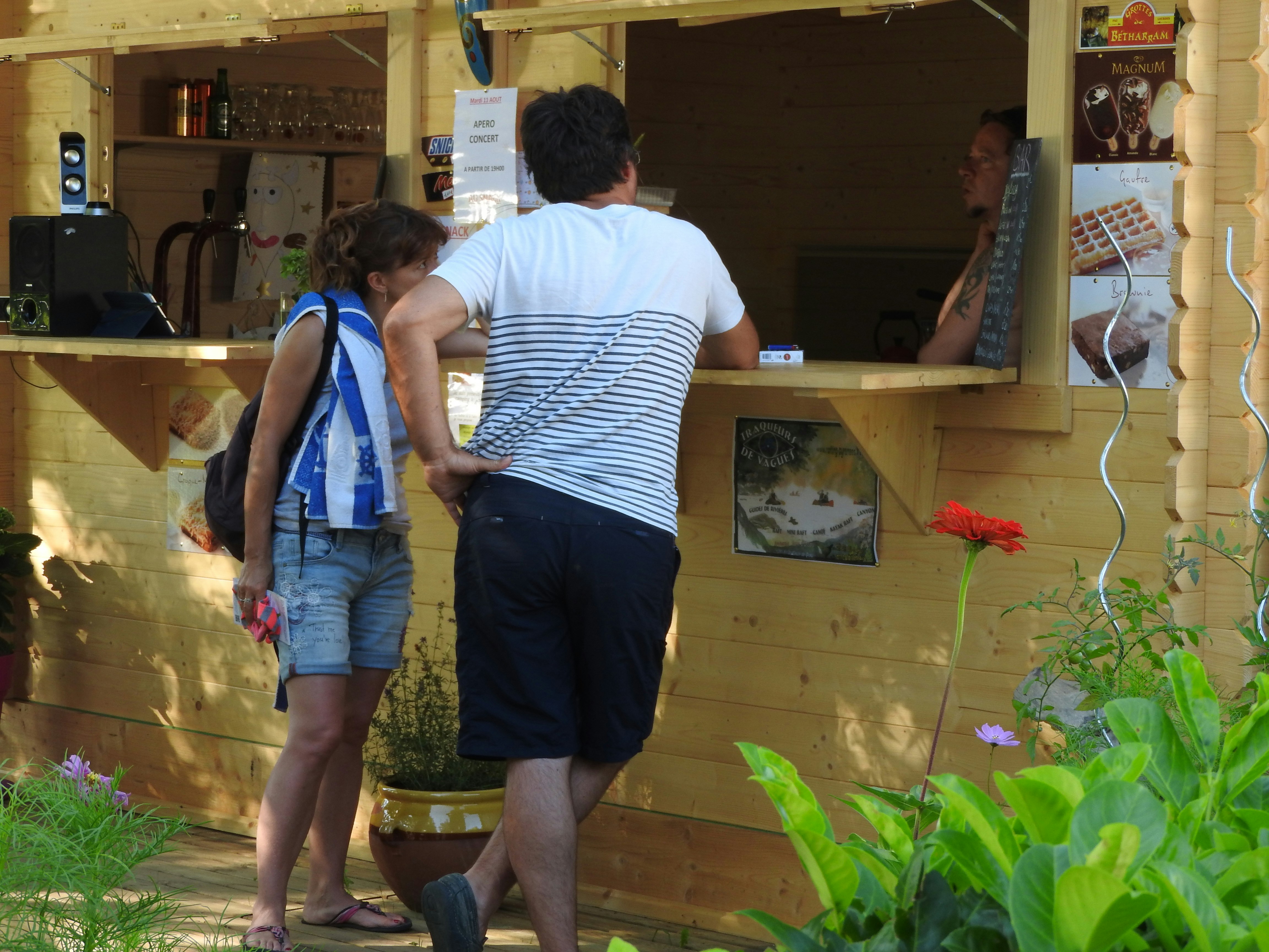 Zwei Personen stehen vor einer Holzhütte am Campingplatz, im Hintergrund Schilder und Regale mit Getränken.