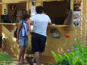 Zwei Personen stehen vor einer Holzhütte am Campingplatz, im Hintergrund Schilder und Regale mit Getränken.