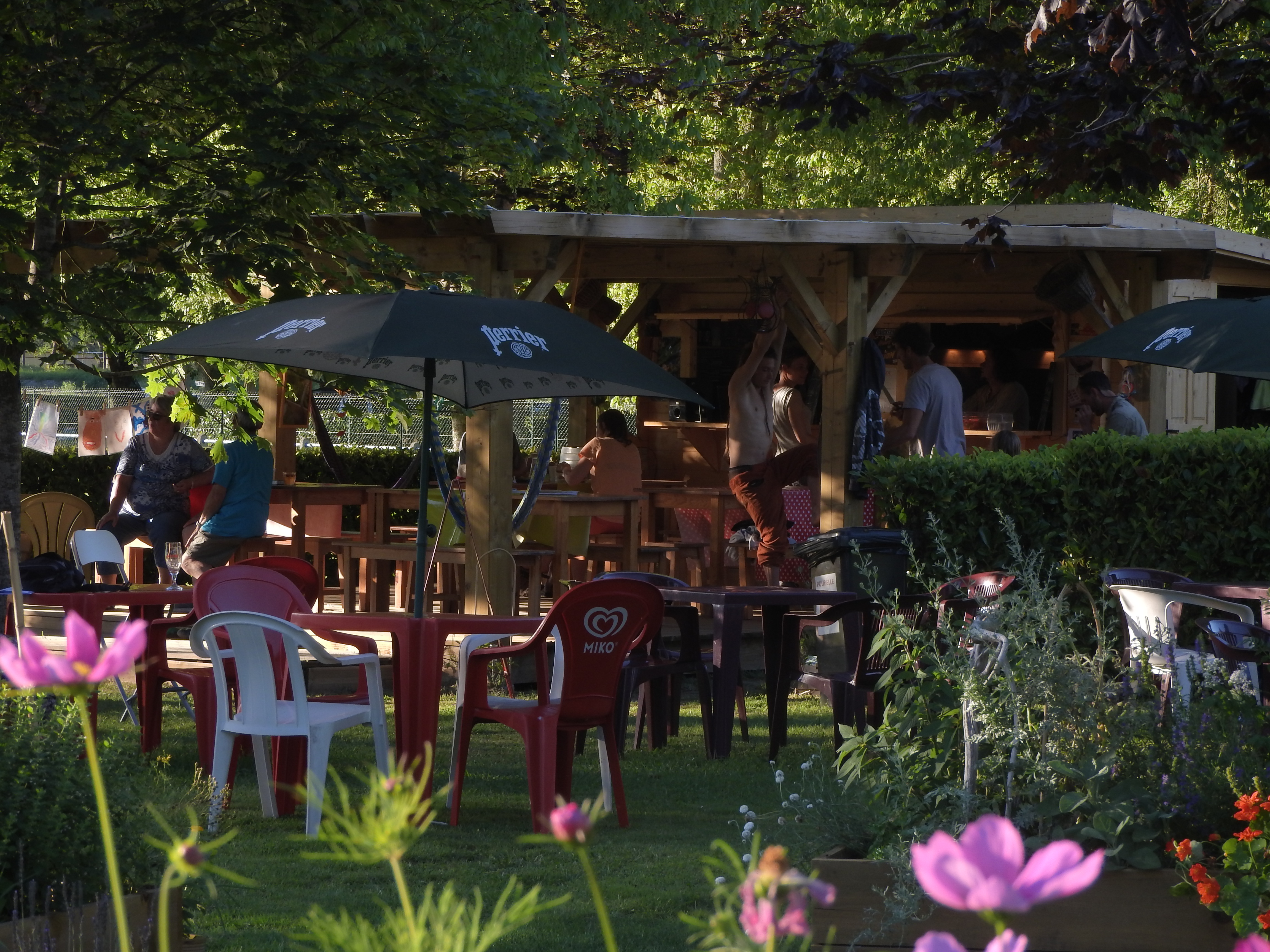 Terrasse am Campingplatz mit Tischen und Stühlen unter Sonnenschirmen, im Hintergrund Bäume und ein Gebäude.