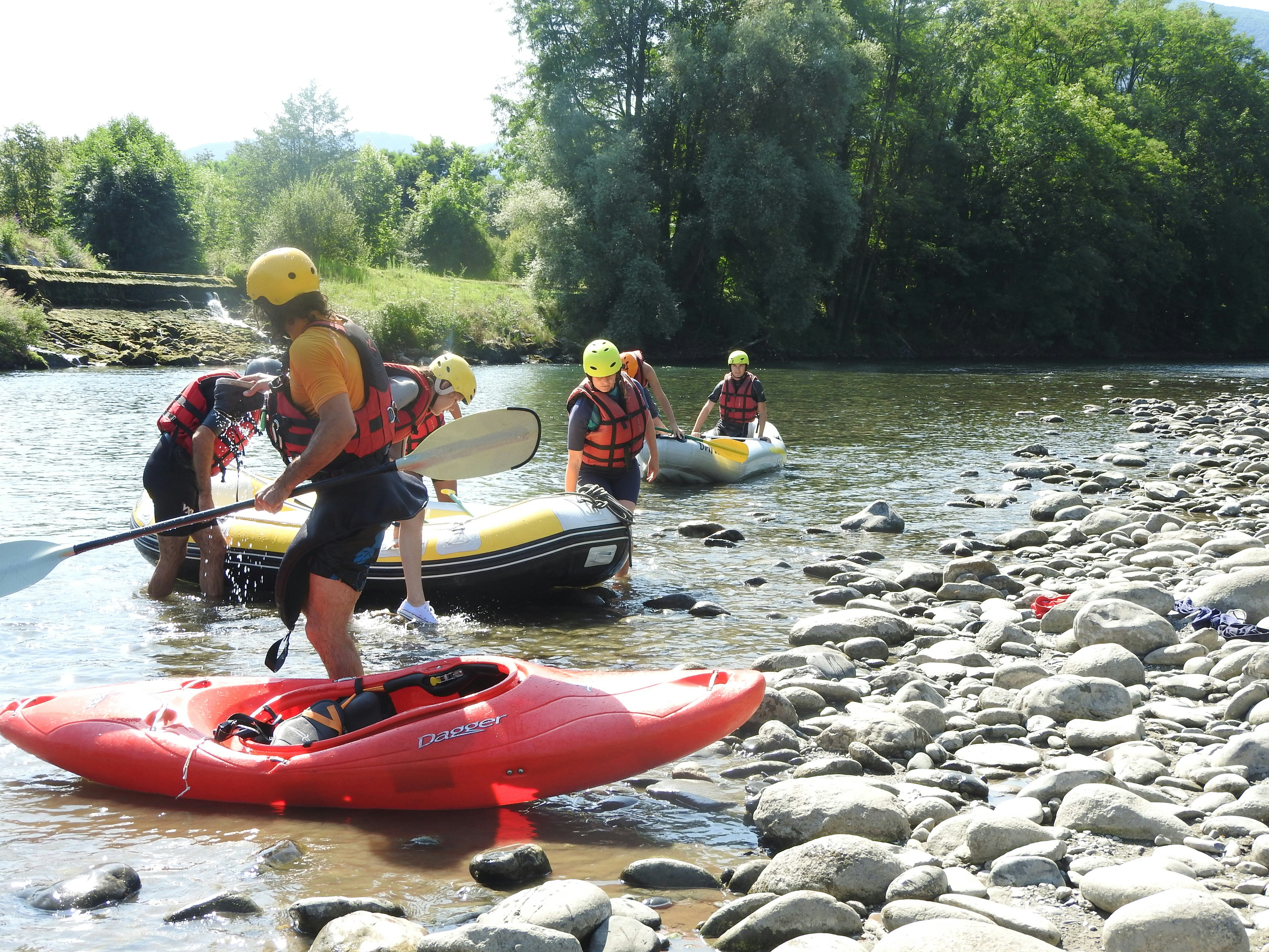 Rafting-Boote auf einem Fluss mit Kieselsteinen am Ufer, mehrere Personen mit Schwimmwesten im Schlauchboot.