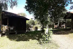 Holz-Campinghäuser mit Terrassen, Garten und Bäumen im Hintergrund, im Vordergrund Rasen und Wege.