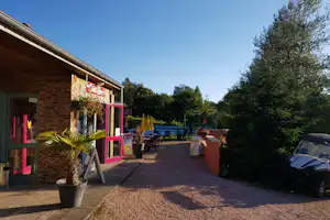 Eingang am Campingplatz mit pinkem Rahmen, Terrasse und Sitzgelegenheiten, daneben Bäume und ein Parkplatz mit Auto.