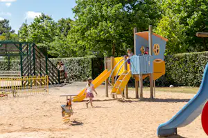 Spielplatz mit gelber Rutsche und Kletterturm im Sand, umgeben von Hecken und Bäumen, im Hintergrund ein Zaun.