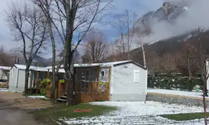 Weißes Mobilheim auf einem Campingplatz im Schnee, mit Holzveranda, Wegen und Bäumen vor den Bergen.