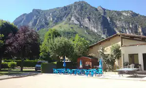 Campinggebäude mit Terrasse, blauen Stühlen und Tischen, umgeben von Bäumen und Blick auf die Berge im Hintergrund.