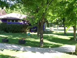 Grüner Campingrasen mit Kiesweg, Bäumen und Blumenkübeln vor einem kleinen Gebäude mit Dachziegeln.
