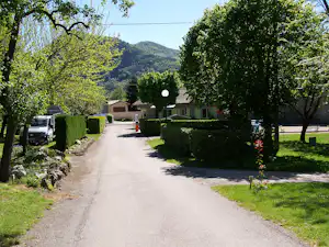 Campingweg zwischen Hecken und kleinen Gebäuden, mit geparktem Wohnmobil und Blick auf grüne Hügel im Hintergrund.