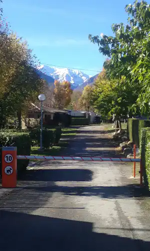 Einfahrt auf einem Campingplatz mit Schranke, Asphaltweg und Bäumen, im Hintergrund Berge und ein Gebäude.