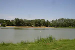 Ruhiger Campingsee mit grünem Ufer, Bäumen und kleinen Booten auf dem Wasser, im Hintergrund Häuser am Rand.