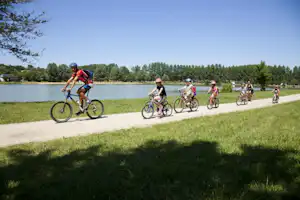 Familie beim Radfahren auf dem Weg am See, mehrere Kinder und Erwachsene mit Fahrrädern, im Hintergrund Bäume und Ufer.